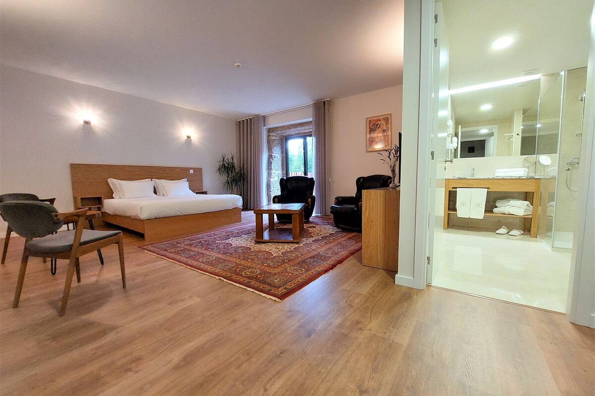 A hotel room with a bed, a desk, a chair, a sofa, a rug, and a bathroom with a mirror and shelves.