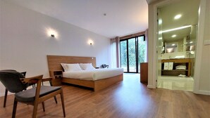A modern bedroom with a wooden bed, a chair, a bedside table, and a bathroom with a mirror and towel rack. - Quinta da Roda - Nature and Wellness (Vila Verde)