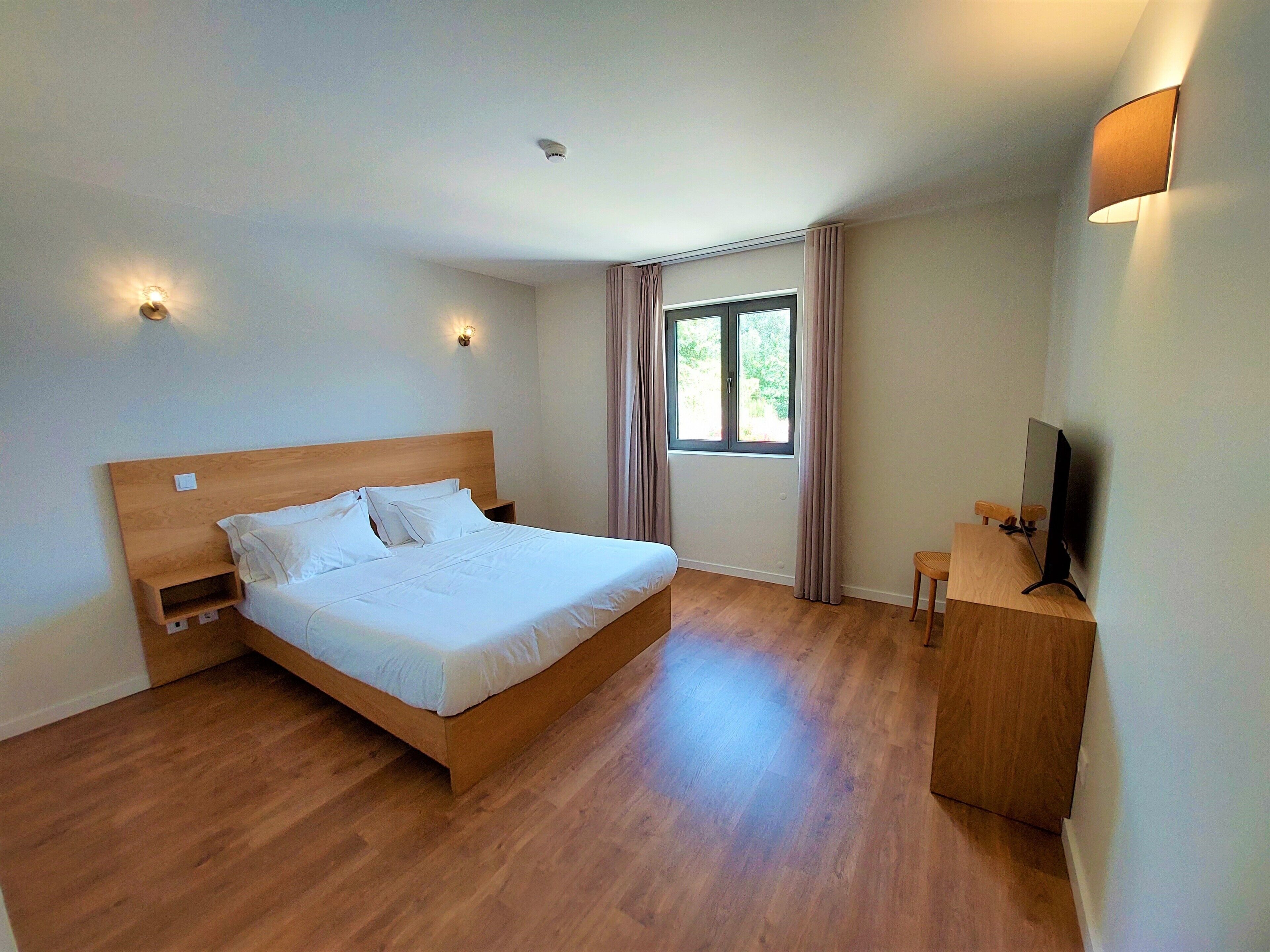 Un dormitorio con una cama de madera, una mesita de noche pequeña, un televisor sobre un mueble de madera y una ventana con cortinas.