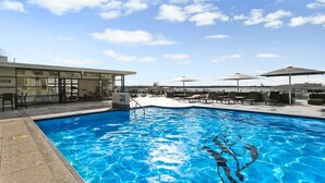 Una piscina con sillones y sombrillas, un edificio con ventanales grandes, y un cielo despejado.