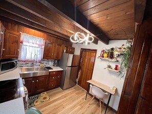 A compact kitchen with wooden cabinets, a stainless steel sink, a microwave, and a refrigerator. There's a small white table with a chair, and a decorative shelf with plants and flowers.