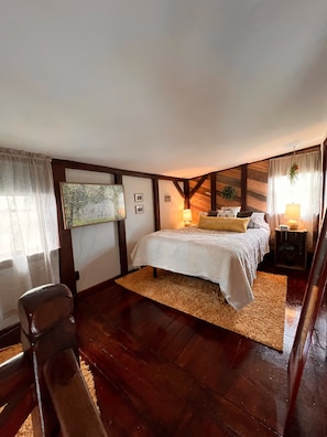 A bedroom with a large bed, wooden beams, a rug, a chair, and a nightstand with a lamp.