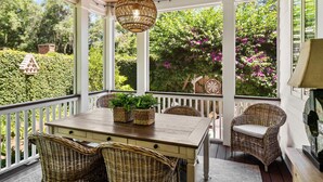 Eine überdachte Veranda mit Holztisch und Korbstühlen, umgeben von weißen Geländern und großen Fenstern mit Blick auf Grünpflanzen und Blumen.