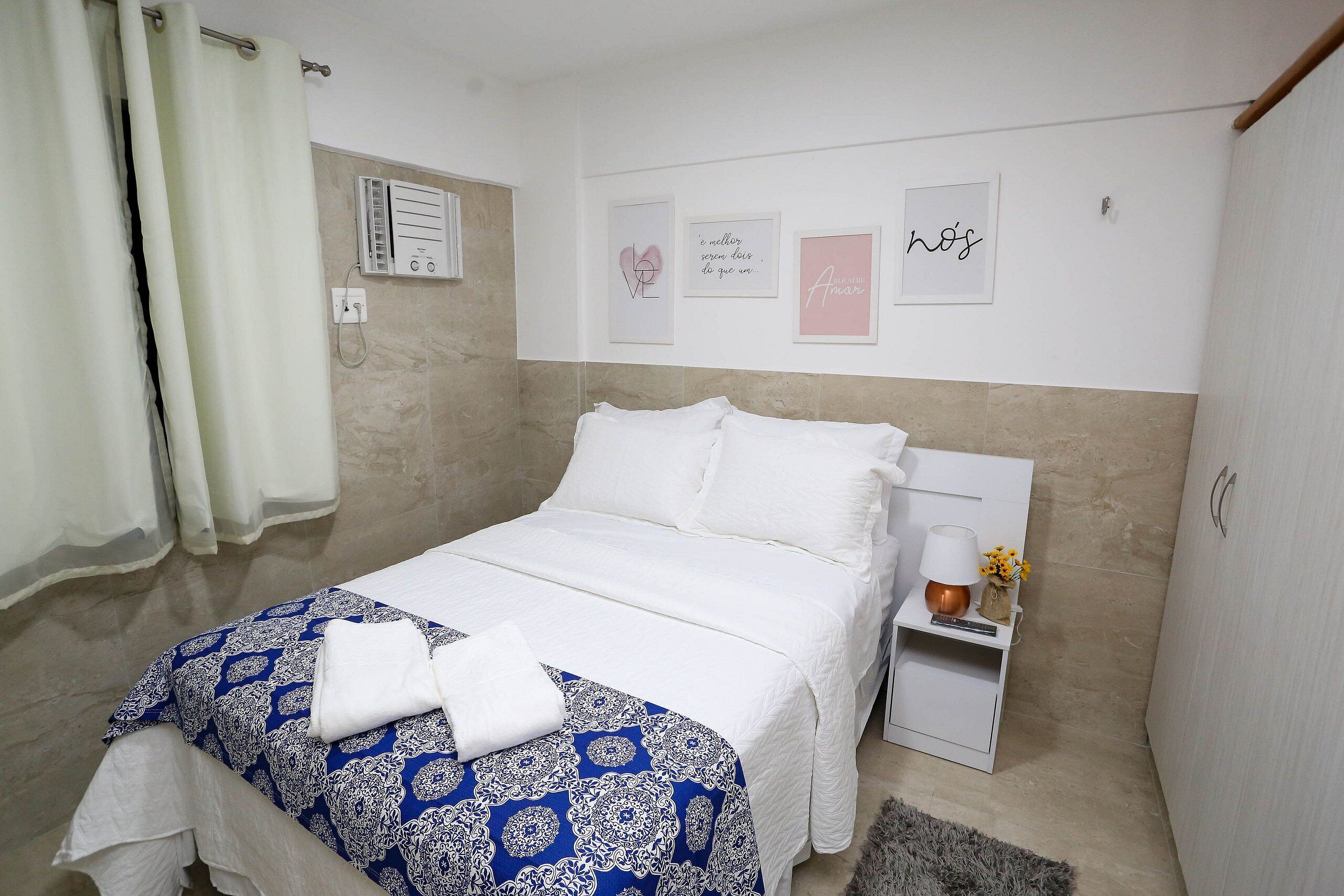 Uma cama bem arrumada com colcha estampada, uma mesa de cabeceira com abajur e um pequeno vaso, e uma parede com quadros de citações inspiradoras.