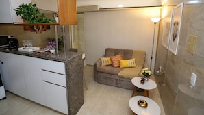 A compact living space with a kitchenette, a sofa with cushions, a small side table, and a decorative plant. - Incrível suíte em Manaíra por Carpediem (João Pessoa)