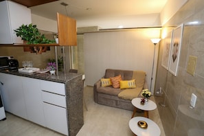 A compact living space with a kitchenette, a sofa with cushions, a small side table, and a decorative plant. - Incrível suíte em Manaíra por Carpediem (João Pessoa)