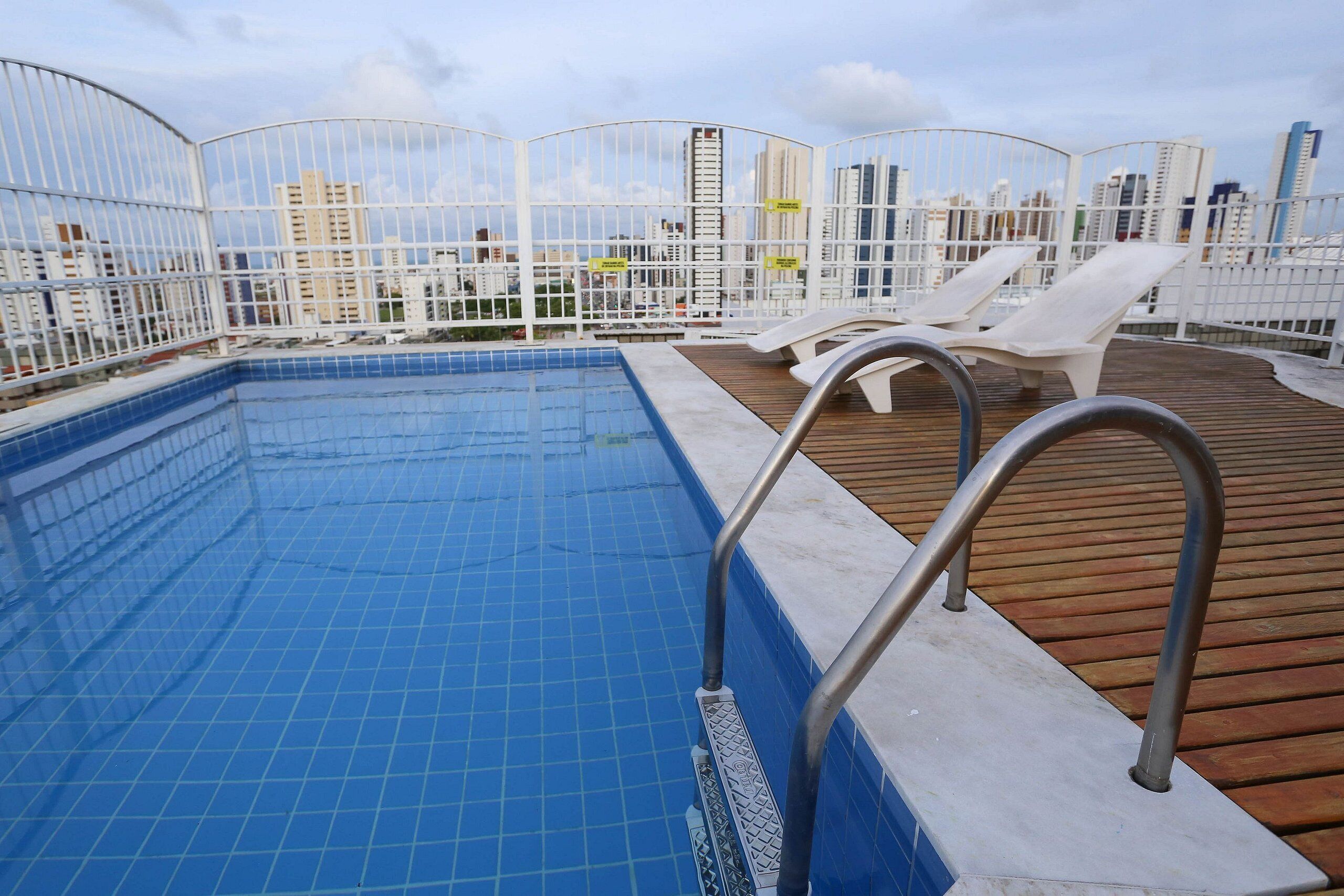 Una piscina en la azotea con azulejos azules, sillas de descanso blancas y una terraza de madera.
