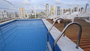 Una piscina en la azotea con azulejos azules, sillas de descanso blancas y una terraza de madera.