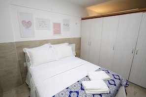 Ein ordentlich bezogenes Bett mit weißen Laken und einer blau gemusterten Tagesdecke, ein Kleiderschrank mit Schiebetüren sowie Wandbilder, darunter ein Herz und motivierende Sprüche.