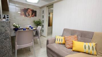 Un salón con un sofá, dos cojines, una mesa de comedor con sillas y una zona de cocina con encimera y una planta en maceta.