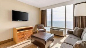 A hotel room with a flat-screen TV mounted on the wall, a sofa, a chair, a wooden side table, and a large window offering a view of the sea.