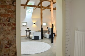 A room with exposed wooden beams, a white brick wall, a round white rug, a sofa, a small table with a plant, and a window with a view of greenery.