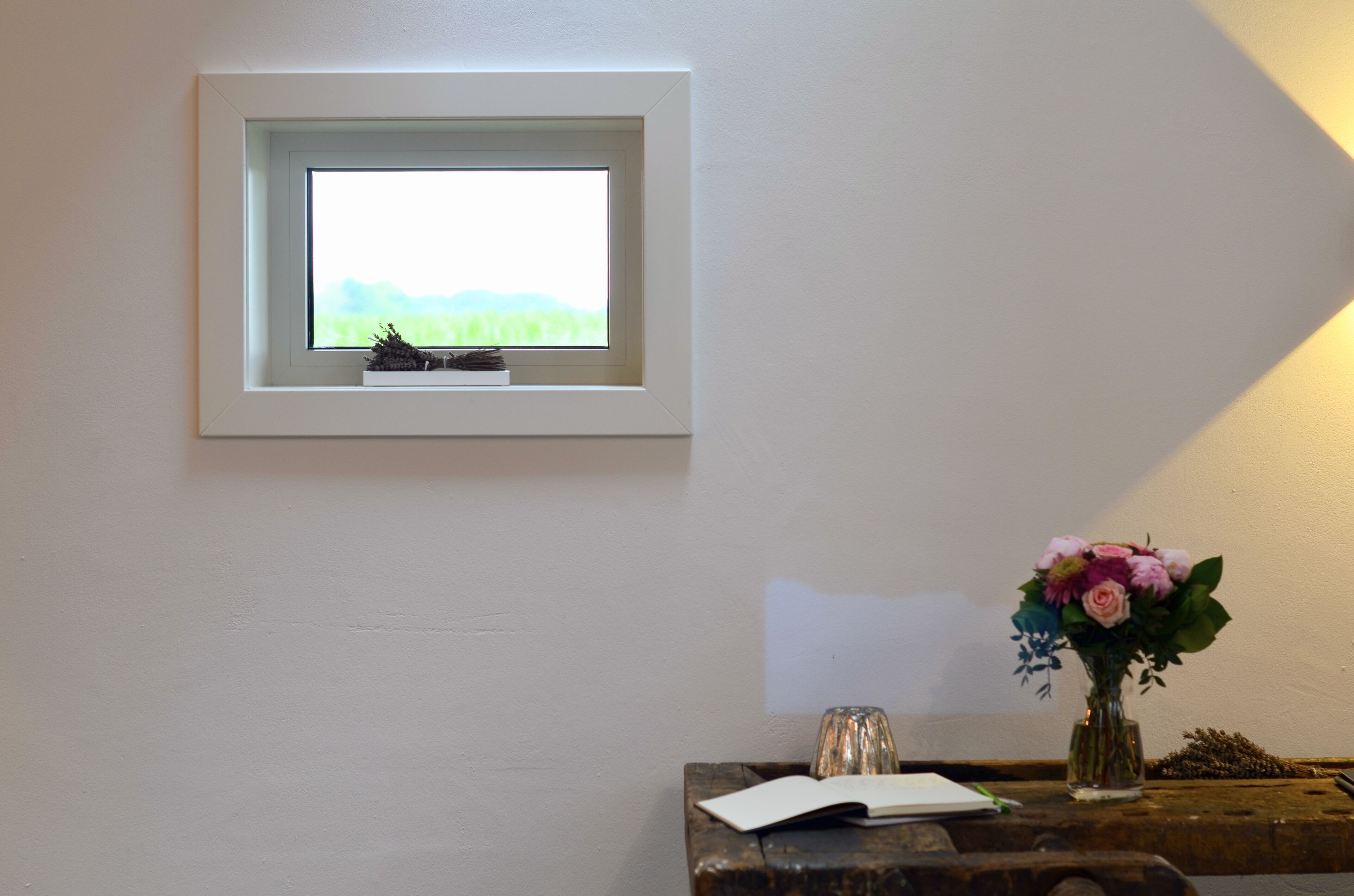 Una ventana pequeña con vistas a la vegetación, un florero con flores y un libro sobre una mesa de madera.