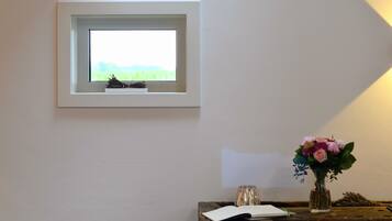 Una ventana pequeña con vistas a la vegetación, un florero con flores y un libro sobre una mesa de madera.