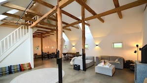 Un loft spacieux avec des poutres apparentes, un escalier, un tapis rond blanc et un coin salon confortable avec un canapé et un pouf.