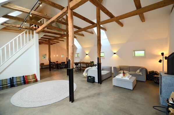 A spacious loft with exposed wooden beams, a staircase, a round white rug, and a cozy seating area with a sofa and ottoman.