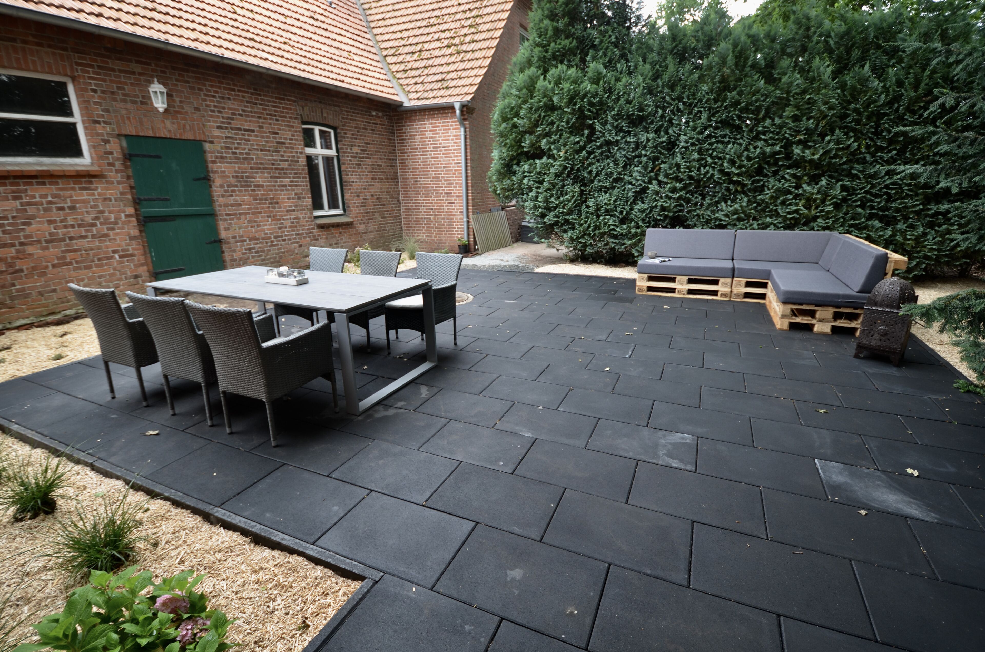 Une terrasse avec une table et des chaises, un banc en bois et, au fond, un bâtiment en briques.