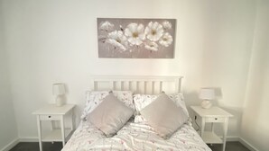 Une chambre à coucher avec un lit blanc, une literie à motifs floraux, deux tables de chevet avec des lampes et un tableau floral accroché au mur.