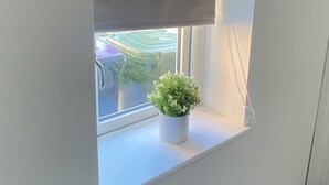 Une petite fenêtre avec un store blanc, une plante en pot et une vue sur un espace extérieur verdoyant.