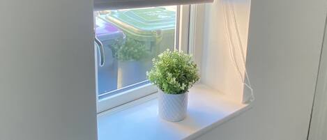 Uma pequena janela com uma persiana branca, um vaso de planta e vista para uma área verde externa.