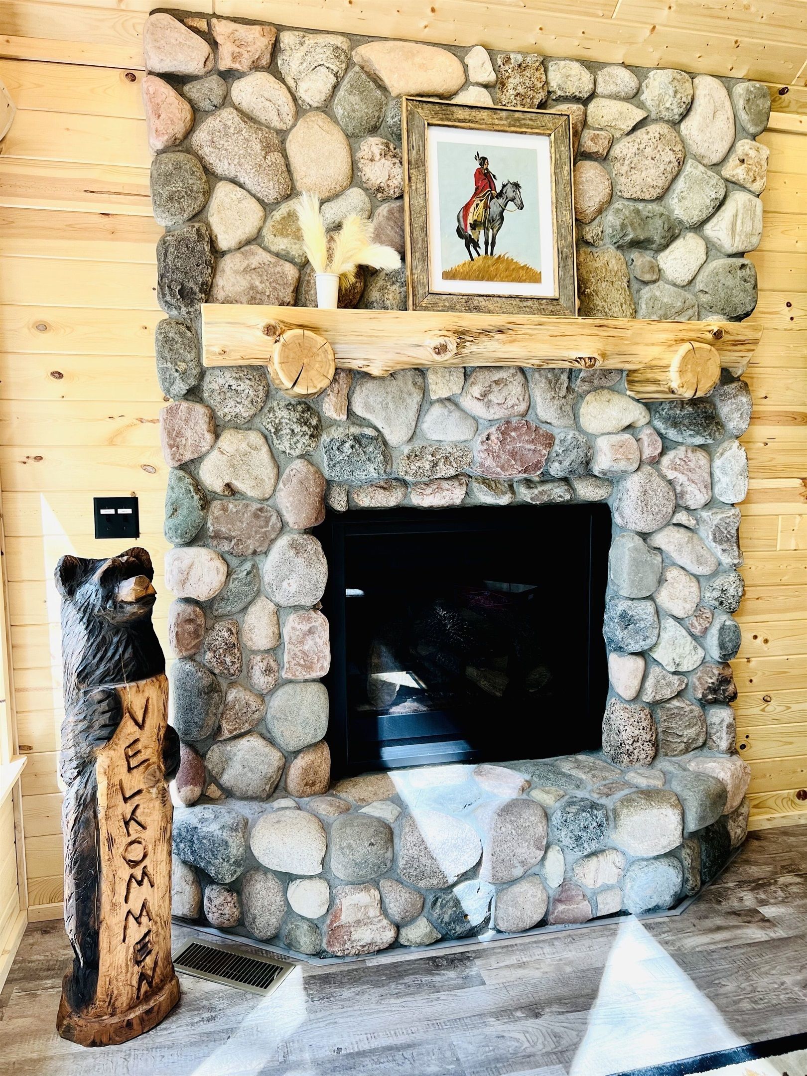 A stone fireplace with a wooden mantel, a framed picture above it, and a bear-shaped wooden sculpture with the word "WELCOME" carved on it.
