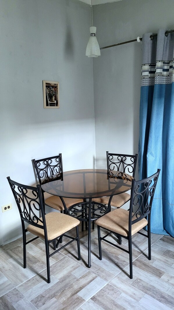 A dining area with a round glass table surrounded by four chairs with cushioned seats and metal frames. There is a hanging light fixture above the table, and a framed picture on the wall.
