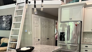 Cocina moderna con armarios blancos, encimera de granito, electrodomésticos de acero inoxidable y un dormitorio tipo altiloce en la parte superior.