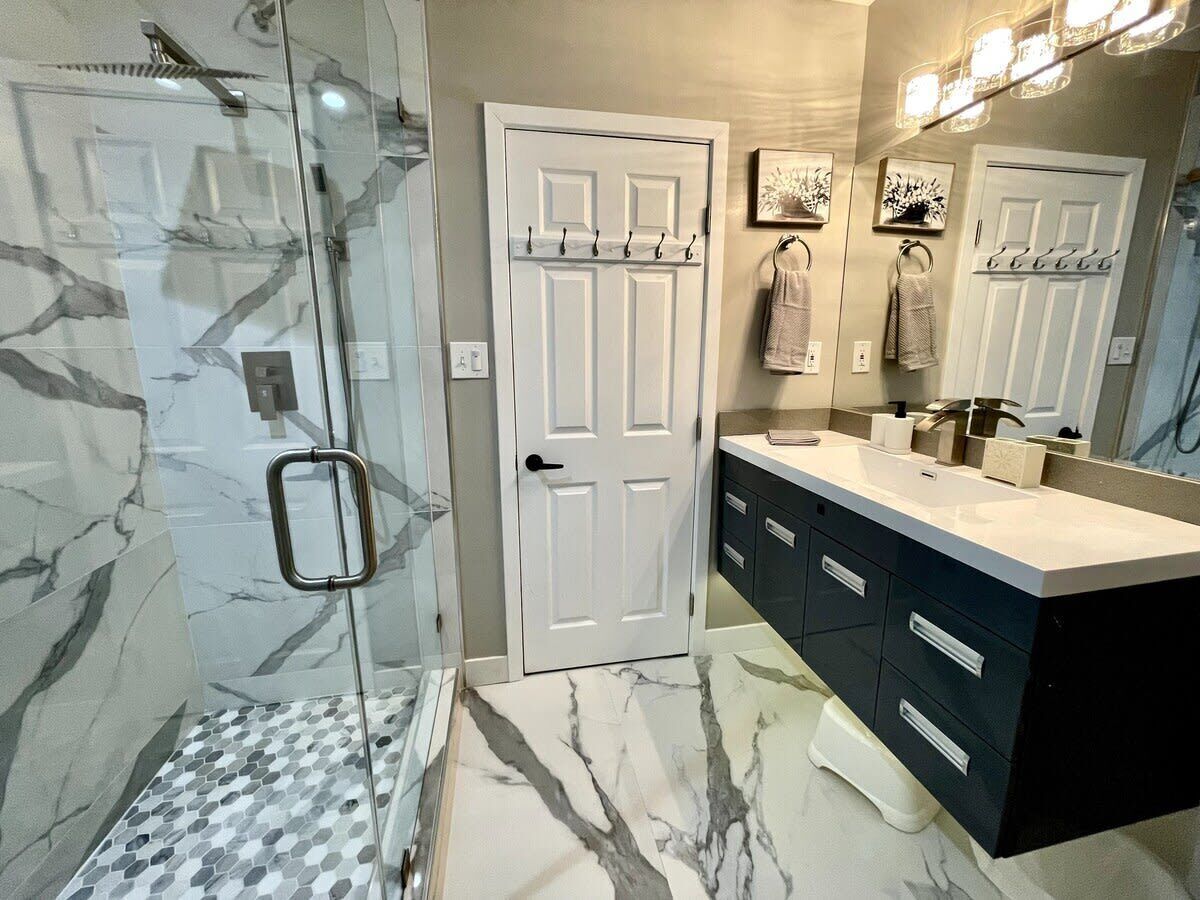 Un baño con cabina de ducha de vidrio, puerta blanca, tocador con armarios oscuros y un espejo grande.