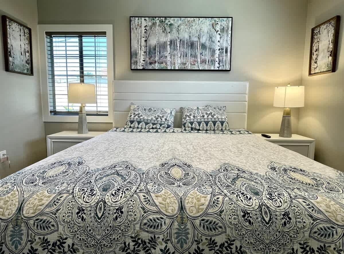 A bedroom with a large bed, two bedside tables, a window with blinds, and wall art.