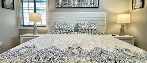 A bedroom with a large bed, two bedside tables, a window with blinds, and wall art.