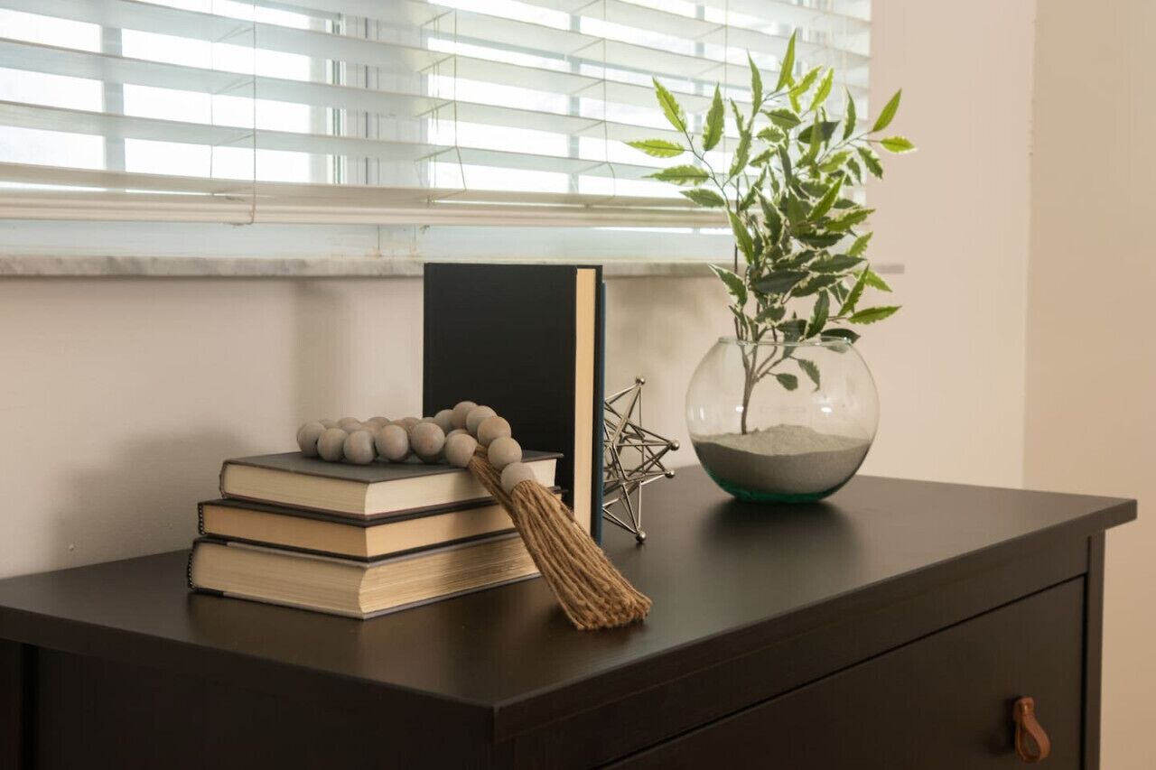 Un escritorio de madera oscura con una pila de libros, un borlón adornado, una escultura geométrica de metal y un florero de vidrio con una planta.