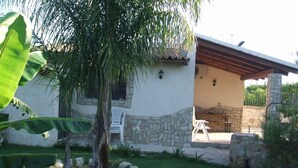 Ein Haus mit einer Fassade aus Stein, einer Terrasse mit Tisch und Stühlen sowie ein kleiner Garten mit Baum und Bananenblättern.