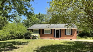 A small, single-story brick house with a dark roof, surrounded by lush green trees and grass. - 3 BR renovated home, near downtown Raleigh, privacy fenced backyard! (Raleigh)