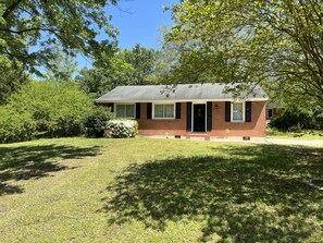 A small, single-story brick house with a dark roof, surrounded by lush green trees and grass.