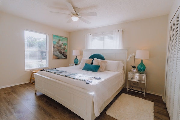 A bedroom with a white bed, a ceiling fan, two bedside tables with lamps, a window with blinds, and a painting on the wall.