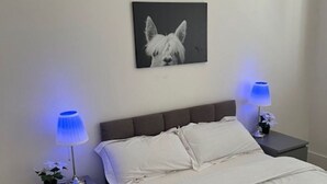 Un dormitorio con una cama, dos mesitas de noche con lámparas, un cuadro colgado en la pared de un perro y una mesilla con una lámpara.
