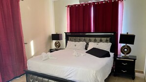 Cama bem arrumada com lençóis brancos e duas almofadas, mesa de cabeceira com abajur e cortina vermelha.