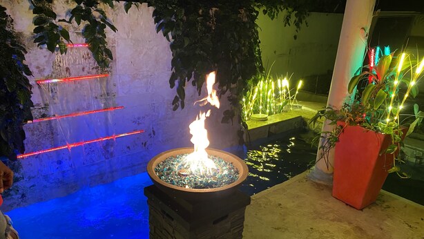 Uma fogueira de pedra com chamas vibrantes, rodeada por um pequeno lago e iluminada por luzes coloridas.