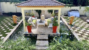 Um gazebo com telhado ladrilhado, rodeado por um jardim quadriculado, um pequeno lago e plantas em vasos.