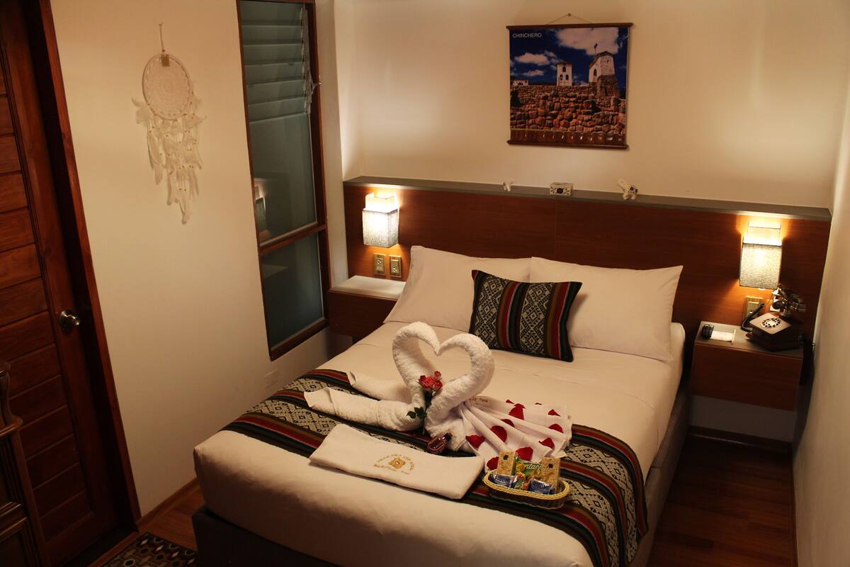 Une chambre d’hôtel avec un lit parfaitement fait, un attrape-rêves accroché au mur, un tableau encadré et une table de chevet avec une lampe et un téléphone.