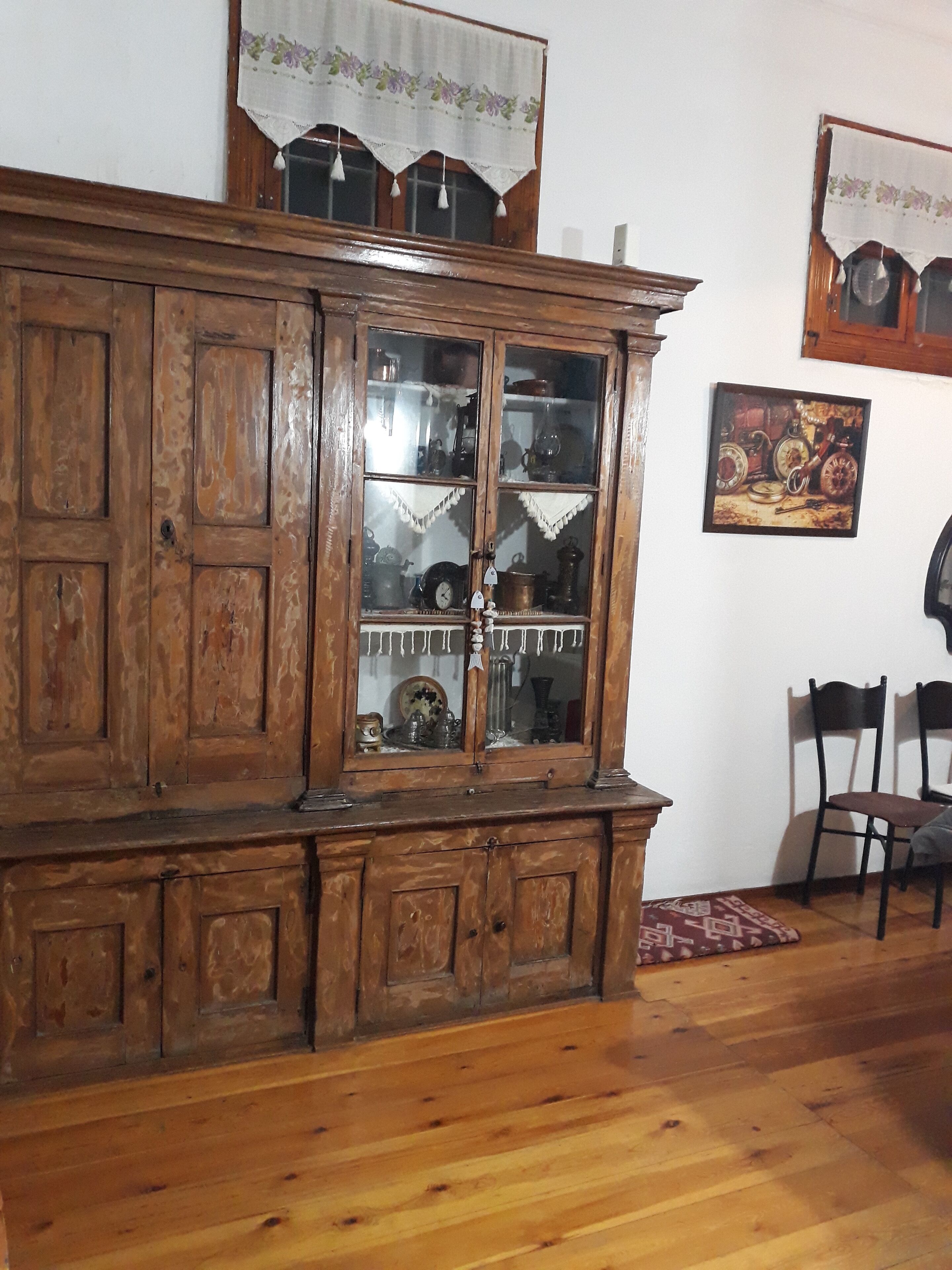 A wooden cabinet with glass doors containing various items. There are two chairs and a small rug on the floor.