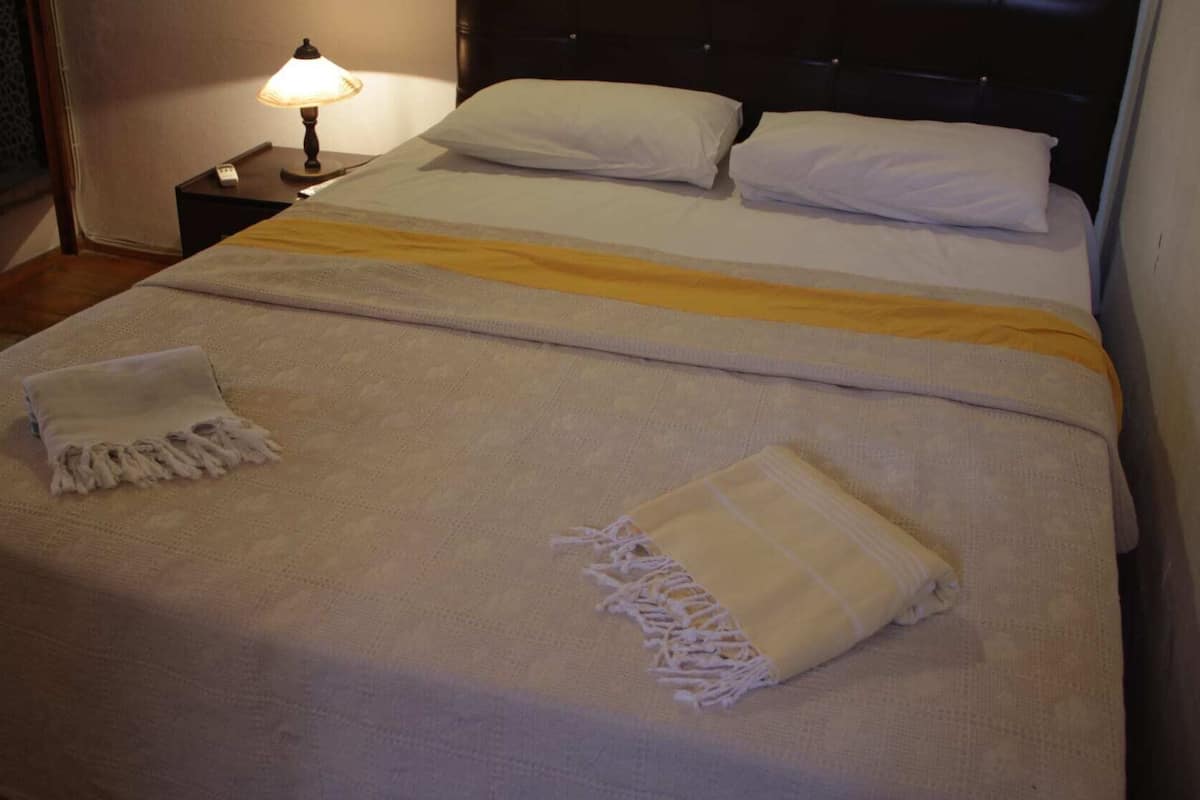 Un lit double avec une couvre-lit blanche et jaune, deux oreillers blancs et une lampe de chevet sur une table de nuit.