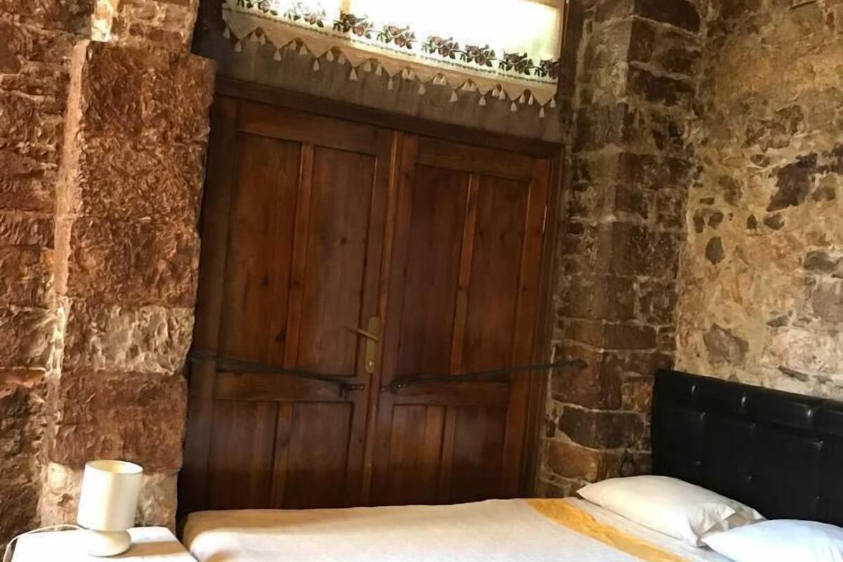 Une chambre avec un mur en pierre, une porte en bois, un lit avec une literie blanche et une table de chevet avec une lampe.