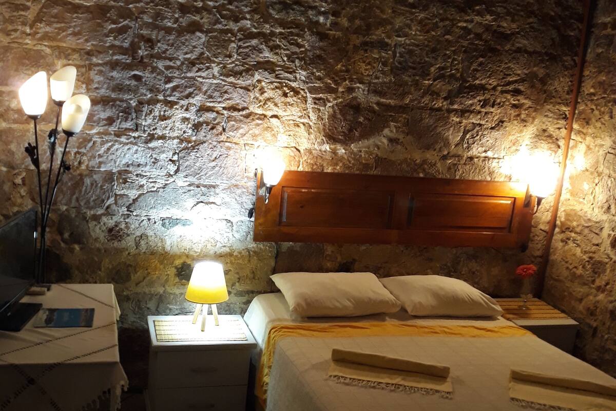 Une pièce rustique aux murs de pierre, comprenant un lit, une table de chevet, une lampe et une chaise.