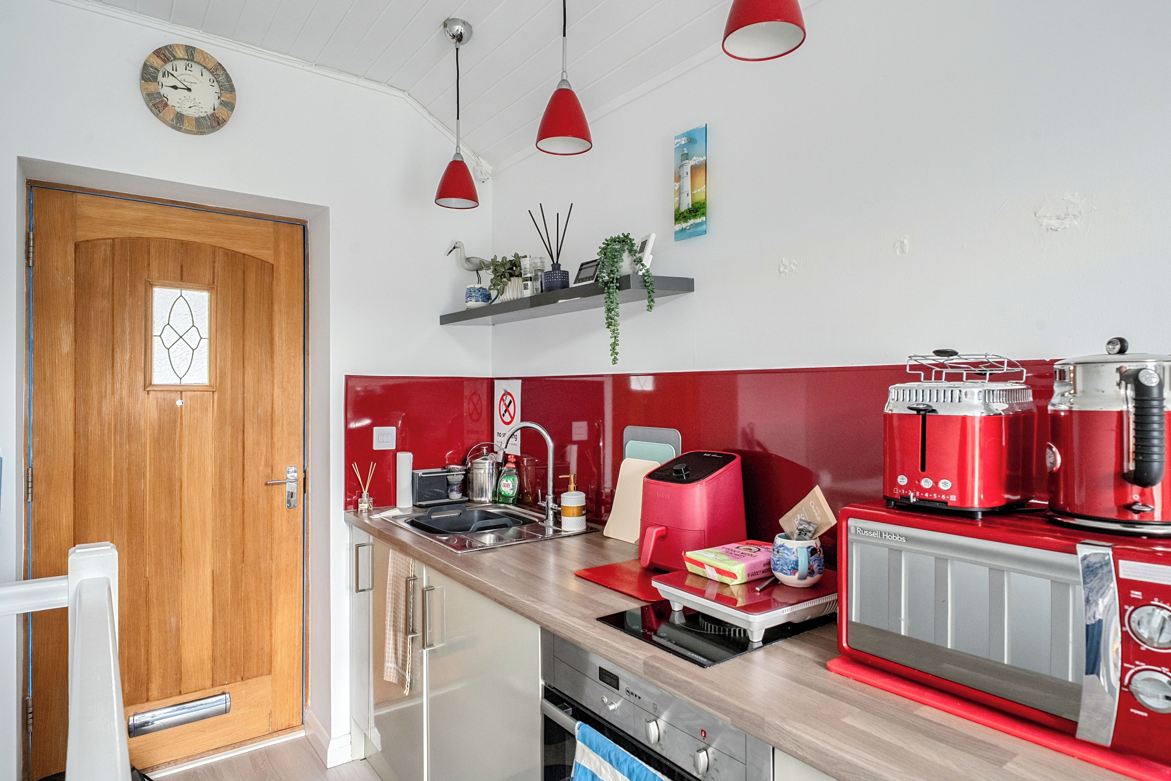 A kitchen with a wooden door, a sink, a red refrigerator, and a clock on the wall.