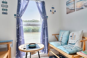 Une pièce avec vue sur un plan d’eau, un canapé agrémenté de coussins sur le thème de la marine, une petite table avec un bol, et un mur décoré sur le thème de la marine.