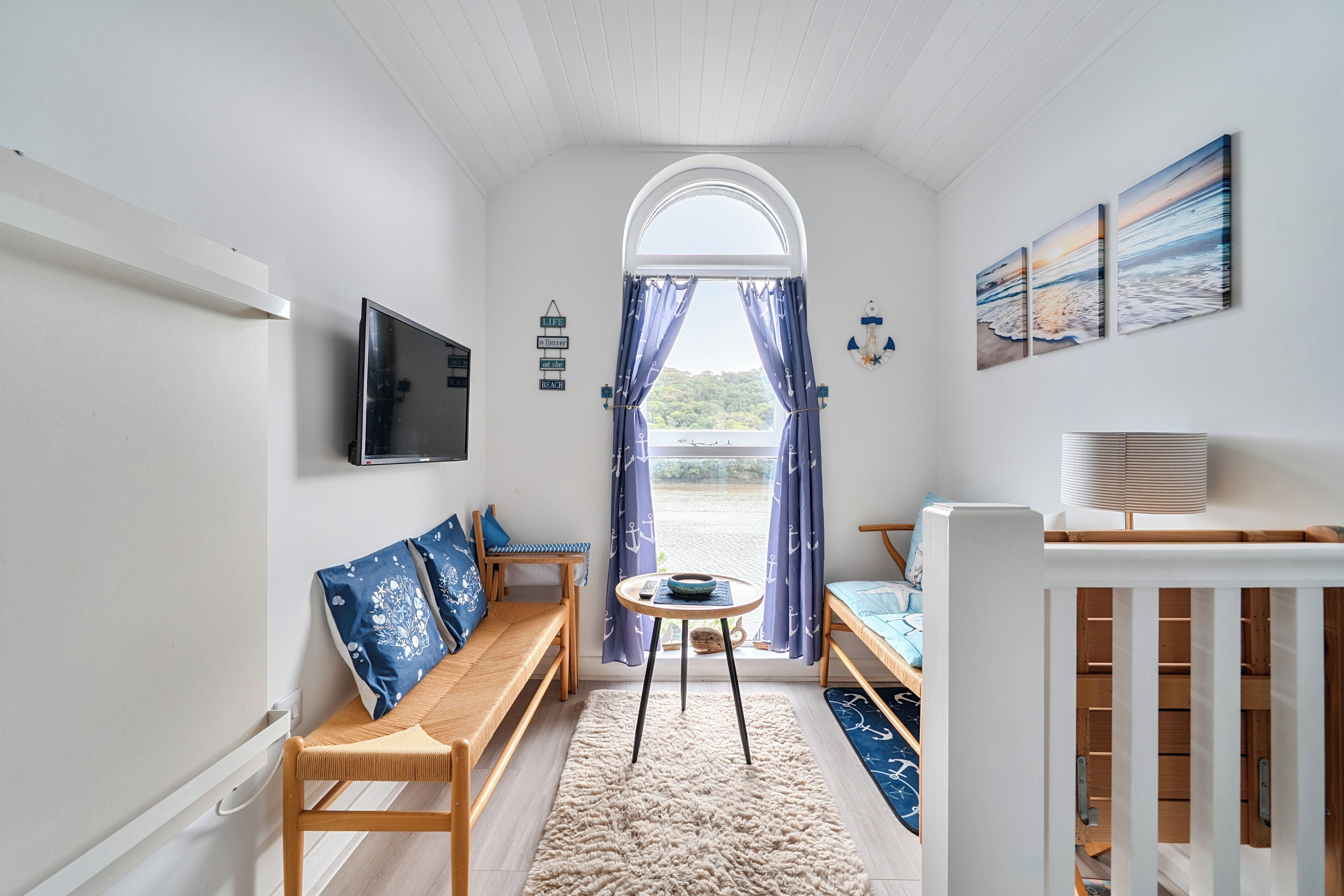Una habitación pequeña con un banco de madera, una mesita redonda, un televisor montado en la pared y vistas a la playa desde una ventana.
