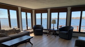 A spacious room with large windows overlooking a body of water, furnished with a sofa set, two armchairs, and a small table with a lamp.