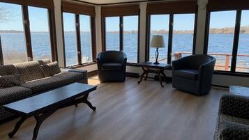 A spacious room with large windows overlooking a body of water, furnished with a sofa set, two armchairs, and a small table with a lamp.