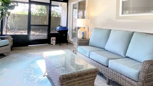A sunlit room with a glass-top table, a light blue sofa, wicker furniture, and a view through large windows.